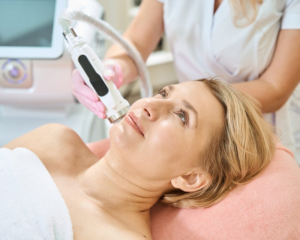 Partial female beautician doing microneedle rf lifting of skin of adult woman Partial female beautician doing microneedle rf lifting of skin with IPL photorejuvenation device of adult european woman in blurred beauty salon. Concept of body skin care and rejuvenation